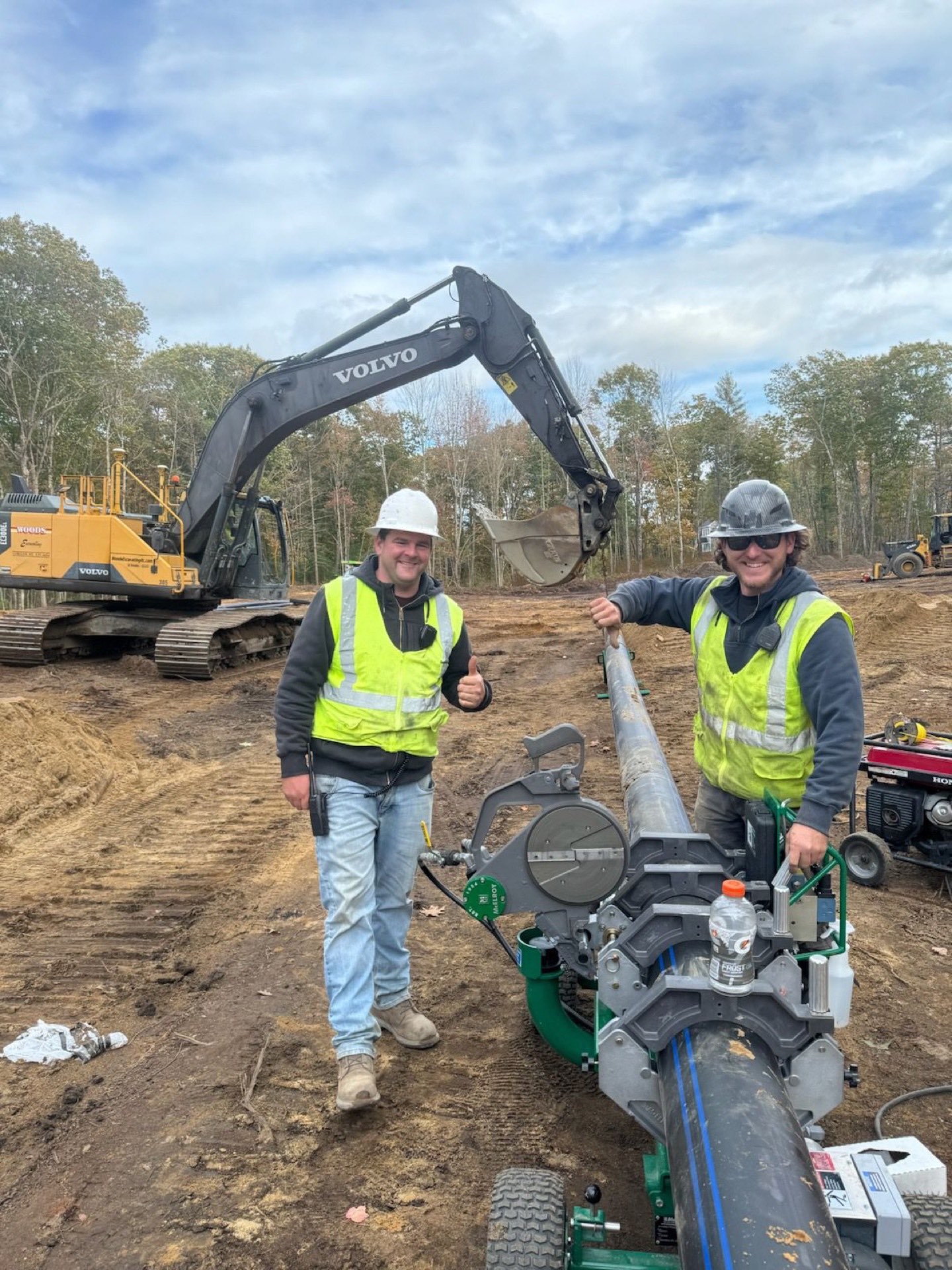 Two smiling Woods crew members in hard hats and hi-vis running a butt-fusion machine on HDPE water main, branded excavator behind