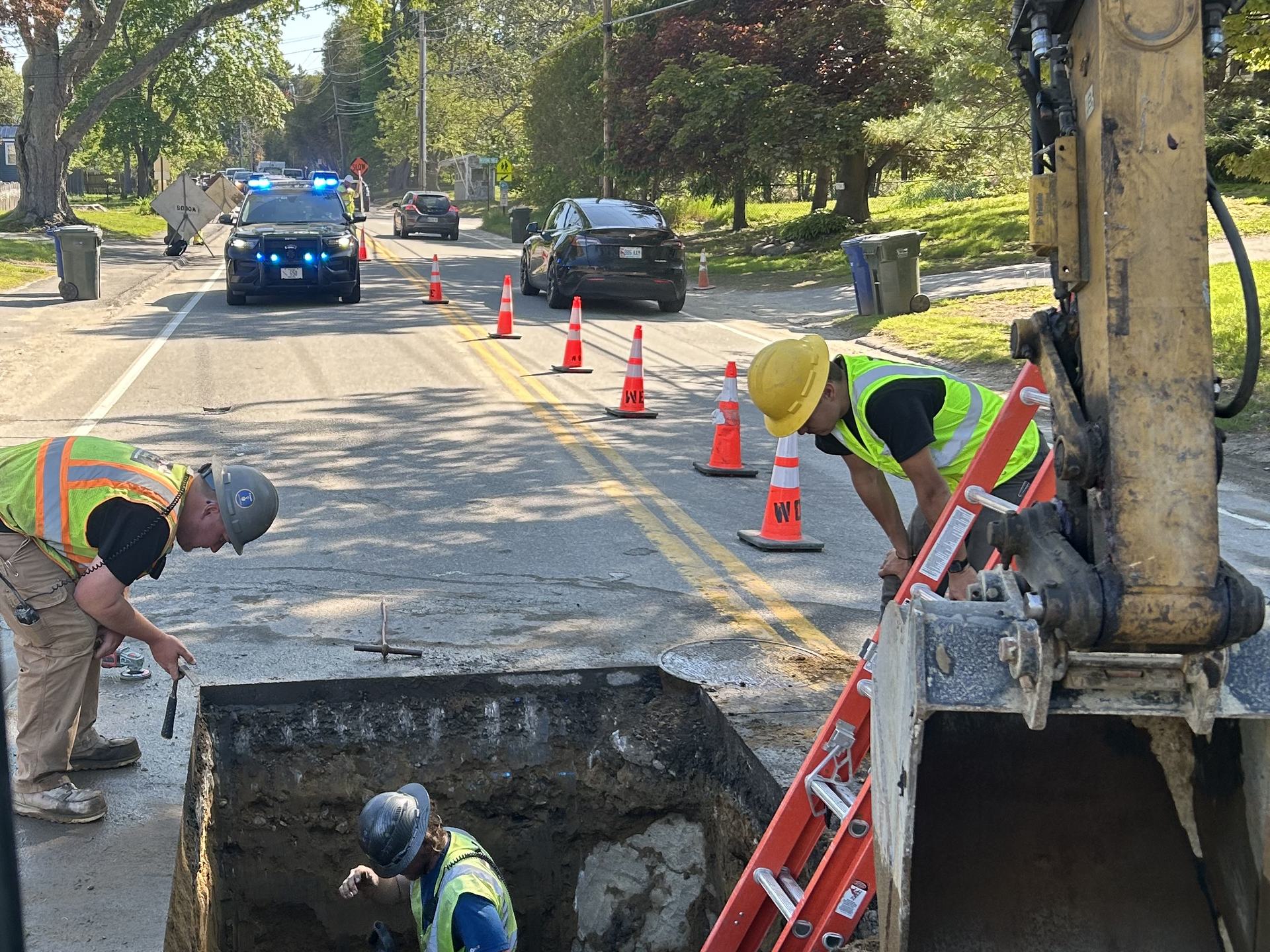 Emergency repair in a live street: crew in a road cut with a ladder set, cones and a police cruiser holding traffic, the excavator bucket at the edge