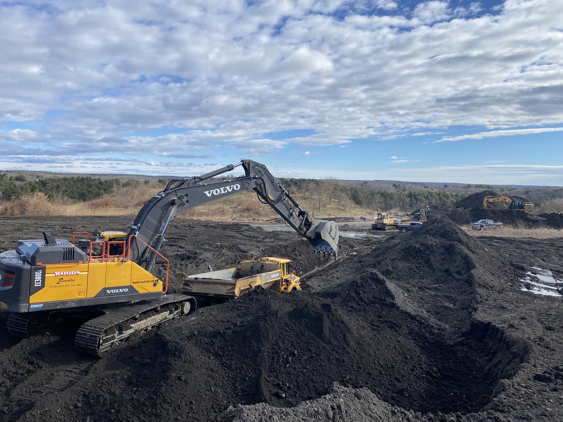 Woods-branded Volvo excavator loading an articulated hauler on a landfill cell, second excavator and dozer behind