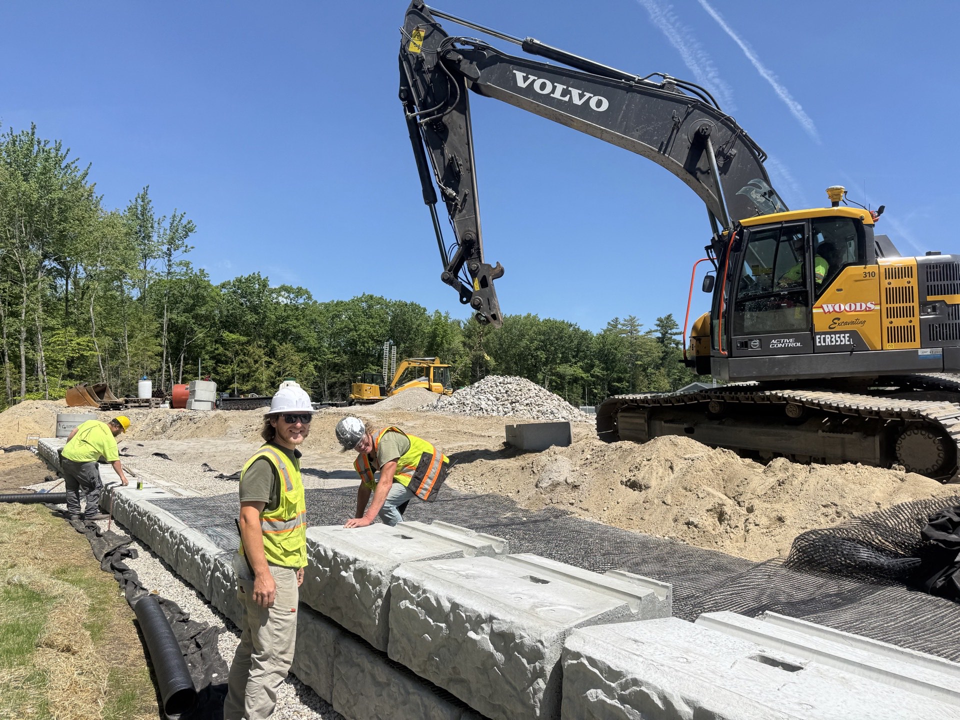 Crew building a precast block retaining wall with geogrid and perforated underdrain, Woods Volvo excavator behind