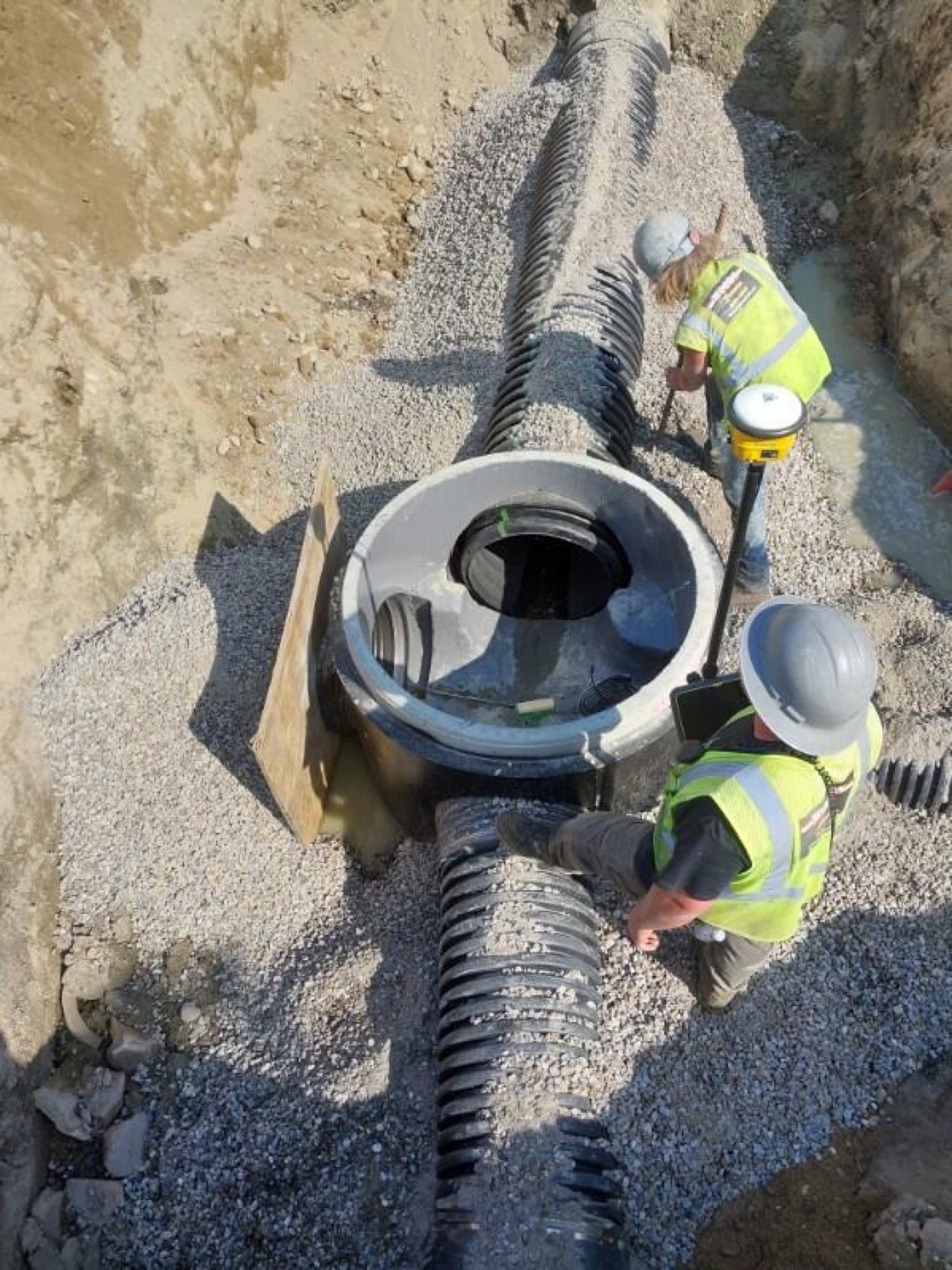 Overhead view of a precast drain structure with HDPE storm pipe on crushed stone bedding, two workers with a GPS rover