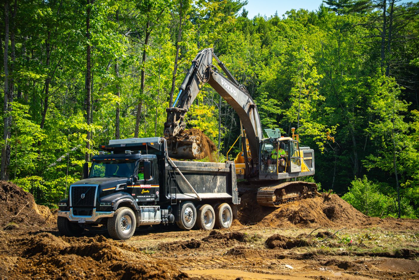 A Woods excavator loading a company tri-axle at a cleared site